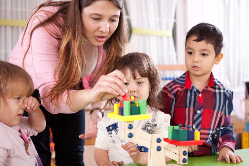 A teacher helping young children balancing some blocks on a scale, perhaps having just give her and the other children proper praise for what they've accomplished.